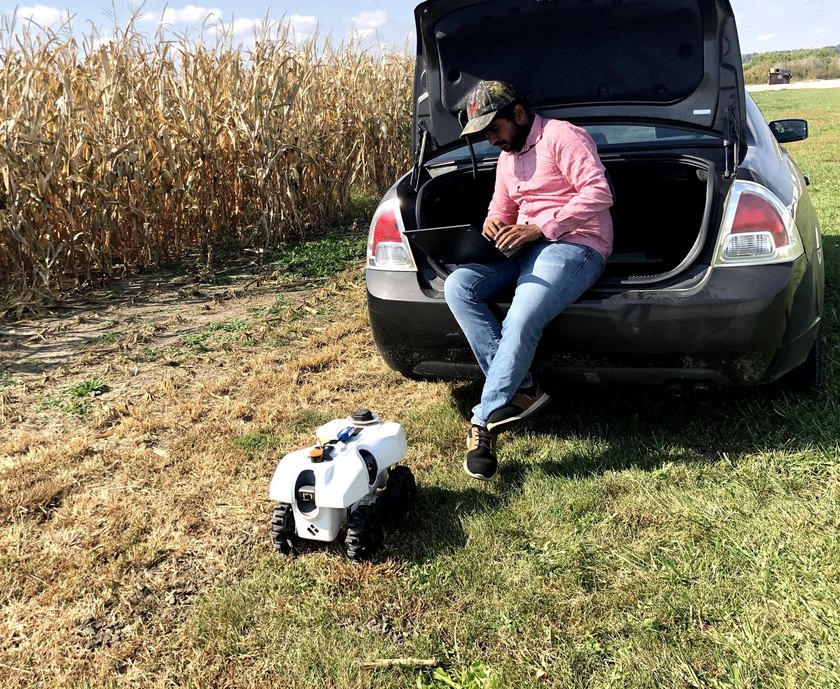 Arun Narenthiran Sivakumar working on a laptop in the open boot of a car at the edge of a corn field, with the robot on the grass alongside.