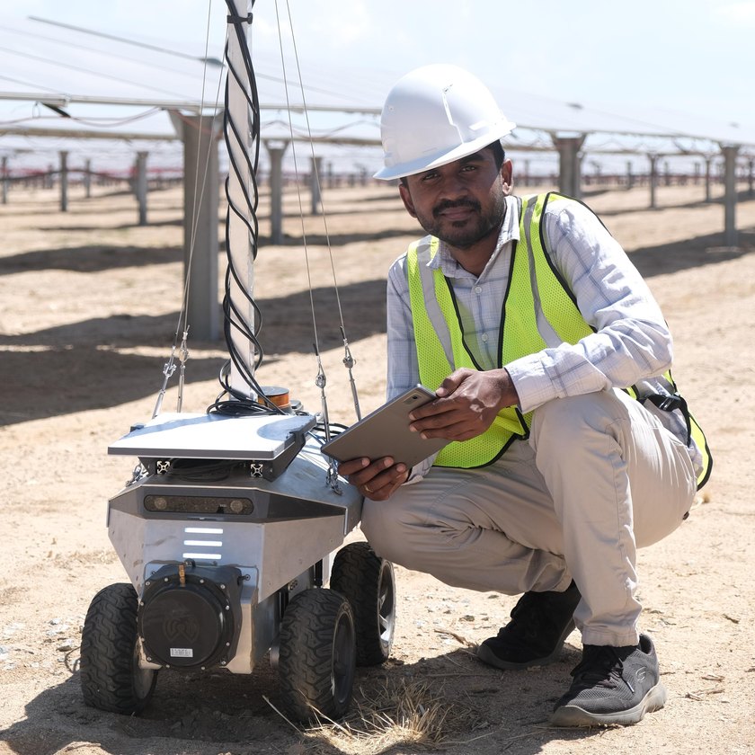 Arun Narenthiran Sivakumar crouched beside the inspection robot on a solar site, in hard hat and hi-vis, holding a tablet.